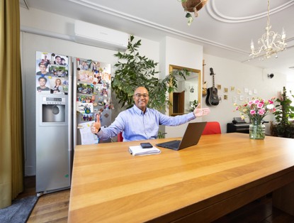 Een persoon in een blauwe blouse zit achter een houten tafel met een laptop en wijst met open armen naar de camera; op de achtergrond zijn een koelkast vol foto's en planten te zien.