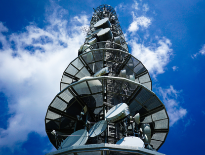 Hoogtelecommast tegen een blauwe lucht met enkele wolken, voorzien van antennes en schotels.