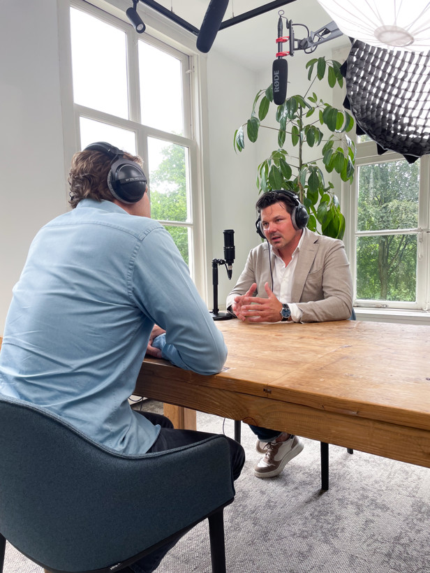 Twee mensen voeren een gesprek aan een houten tafel, omringd door microfoons en opnameapparatuur in een lichte kamer.