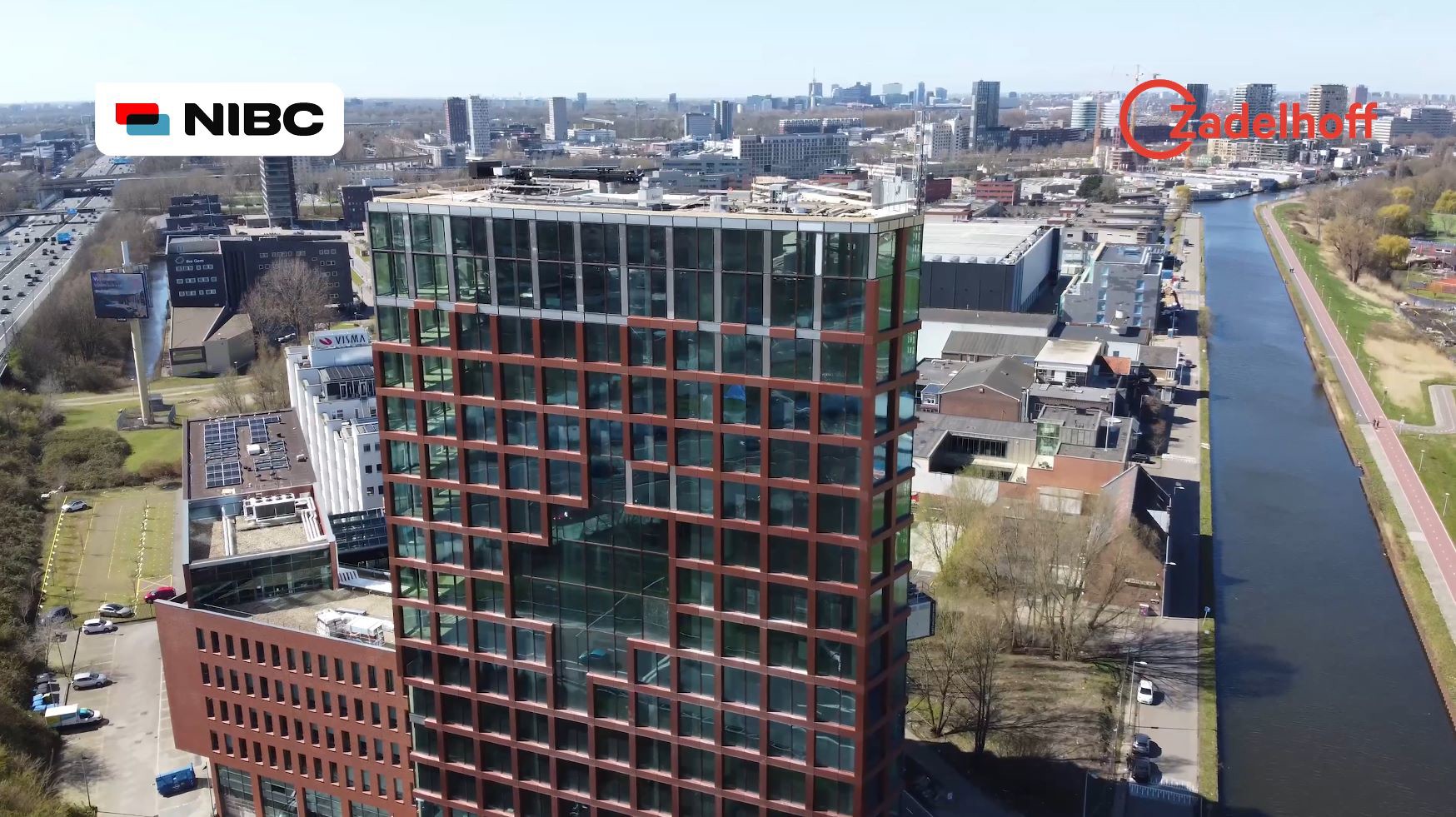 Luchtfoto van een modern kantoorgebouw aan het water, omringd door andere gebouwen en een weg. Het gebouw heeft een glazen façade en is goed zichtbaar.