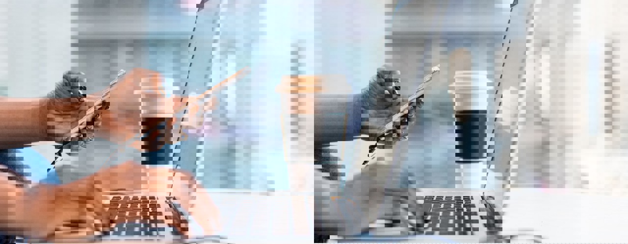 Een persoon met zwart gelakte nagels typt op een laptop terwijl hij/zij een smartphone vasthoudt, met een koffiebeker naast zich.