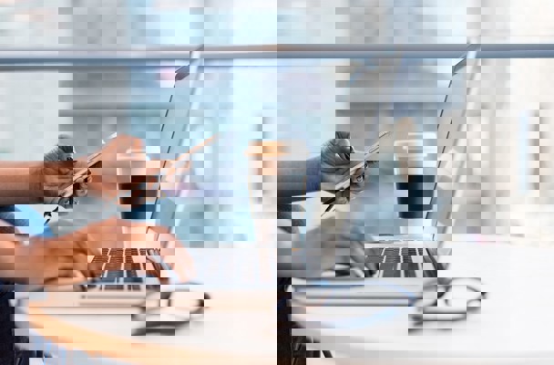 Een persoon met zwart gelakte nagels typt op een laptop terwijl hij/zij een smartphone vasthoudt, met een koffiebeker naast zich.