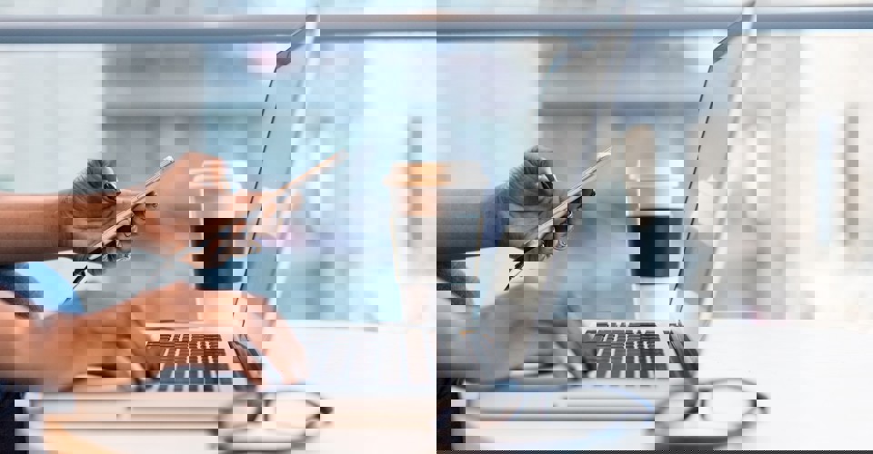 Een persoon met zwart gelakte nagels typt op een laptop terwijl hij/zij een smartphone vasthoudt, met een koffiebeker naast zich.
