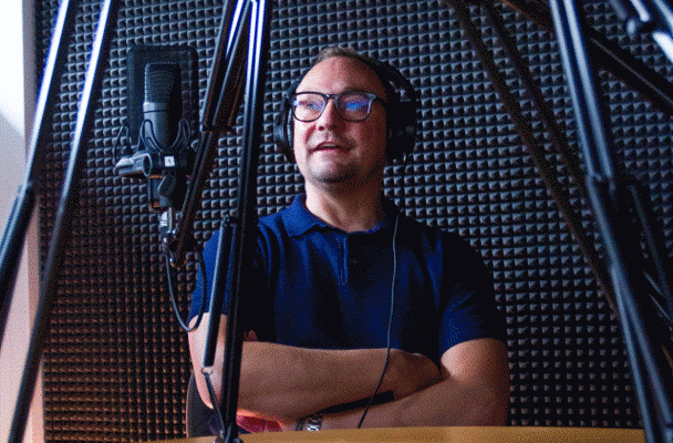 Een persoon met een hoofdtelefoon zit in een geluidsstudio, omringd door microfoons en geluidsisolatiepanelen.