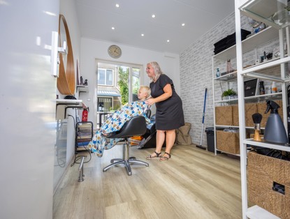 Kapper assisteert een oudere klant in een kleurrijke cape tijdens een haarbehandeling in een lichte, moderne salon.