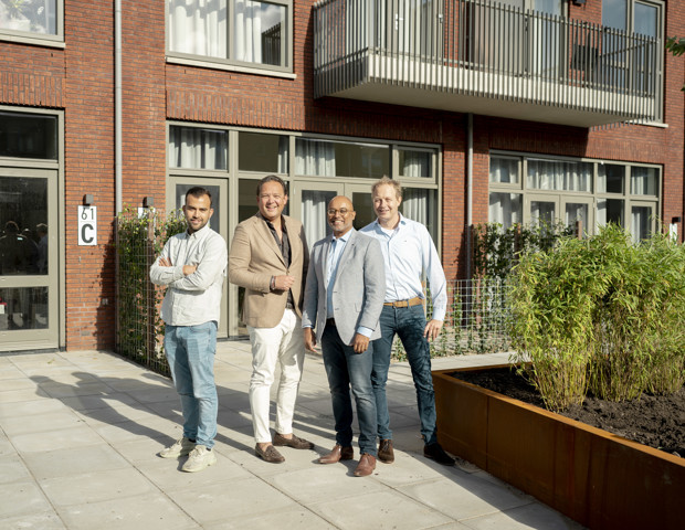 Groep van vier mannen in formele kleding voor een modern appartementencomplex met een groene plantenbak.