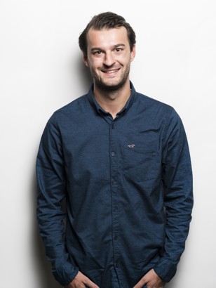 Man in een blauw overhemd staat rechtop met zijn handen in zijn zakken, voor een witte achtergrond.