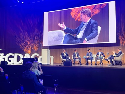 Paneldiscussie op een podium met vier sprekers, met de hashtag #GDAC op de voorgrond en een luisterend publiek.