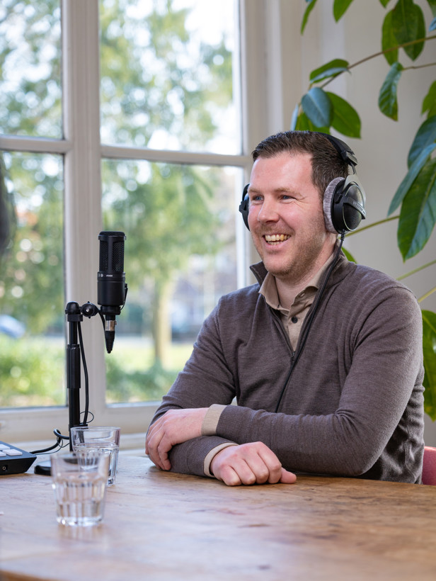 Sven met een microfoon en koptelefoon, leunend tegen een raam met groene planten op de achtergrond.