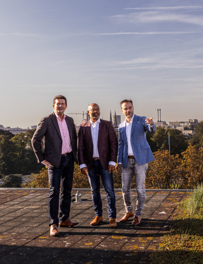 Drie mannen in formele kleding staan op een dak met uitzicht op een stadslandschap en groen in de achtergrond.