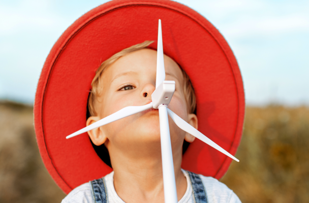 Een kind met een grote rode hoed houdt een plastic windmolen vast in een groene buitenomgeving.