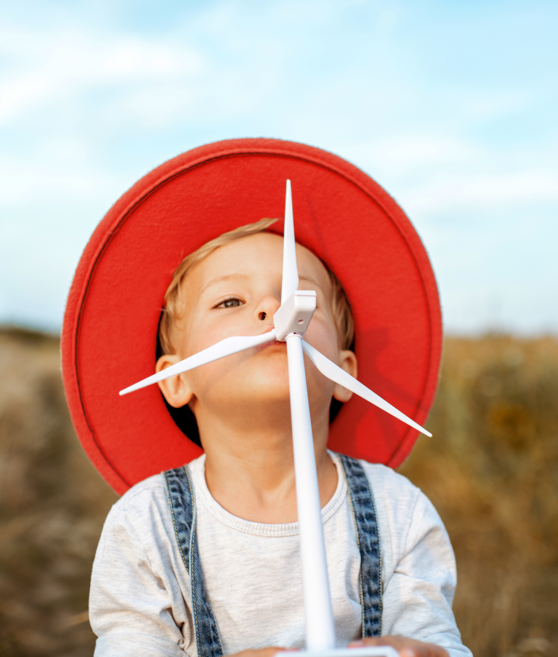 Een kind met een grote rode hoed houdt een plastic windmolen vast in een groene buitenomgeving.