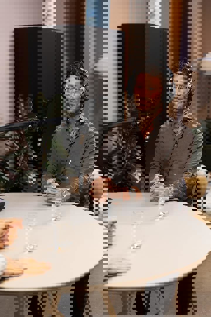 Een persoon met een hoofdtelefoon zit aan een tafel met een microfoon naar hem gericht. Op de tafel staan twee glazen water.