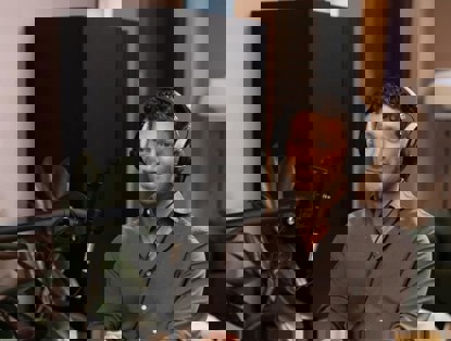 Een persoon met een hoofdtelefoon zit aan een tafel met een microfoon naar hem gericht. Op de tafel staan twee glazen water.