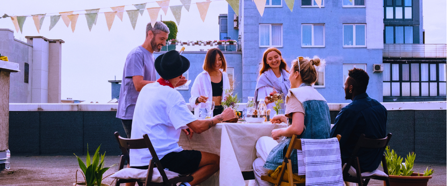 A group of six people sitting at a table on a rooftop terrace, enjoying a pleasant meal and conversations, surrounded by plants and flags.