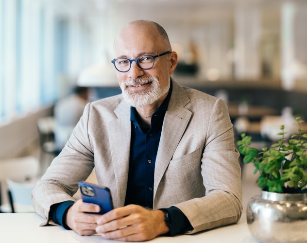 Een man in een lichte blazer zit aan een tafel met een smartphone in zijn hand, omgeven door een moderne interieur.