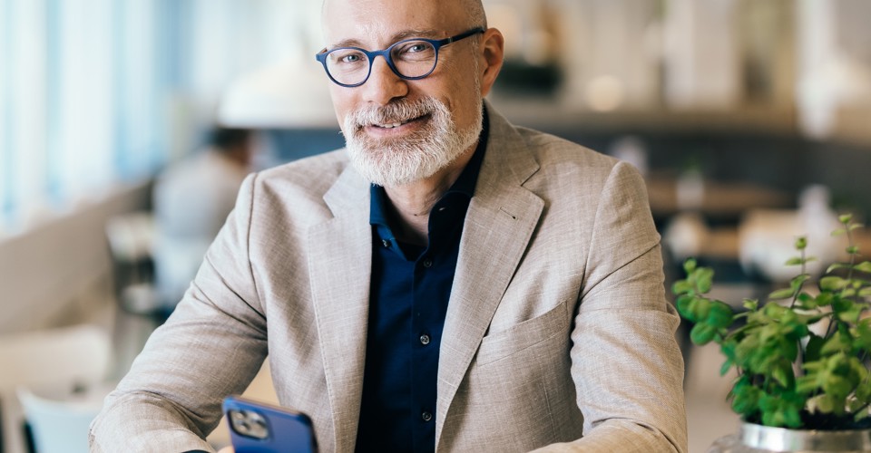 Een man in een lichte blazer zit aan een tafel met een smartphone in zijn hand, omgeven door een moderne interieur.