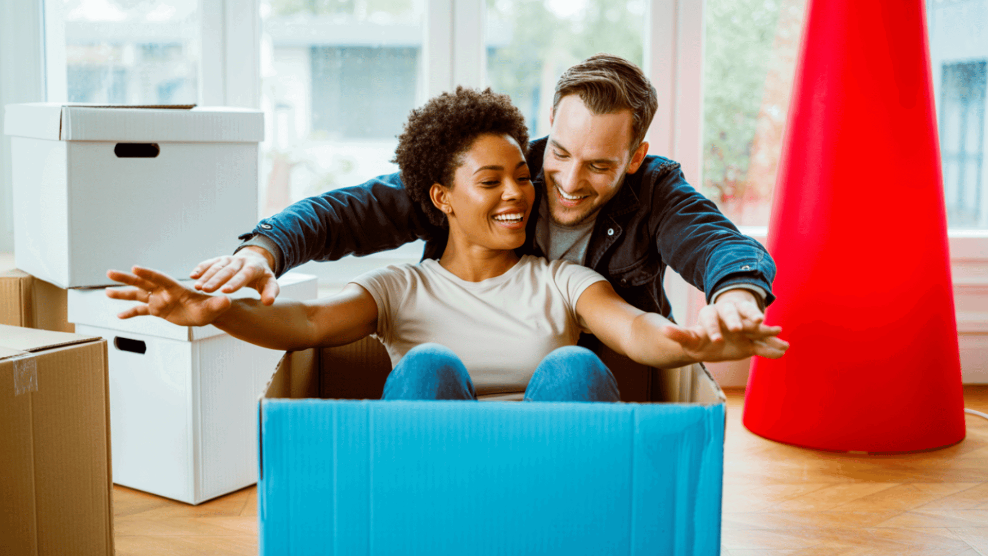 Een jongeman en jongedame zitten met uitgestrekte armen in een grote blauwe kartonnen doos, omringd door verhuisdozen, en hebben plezier.