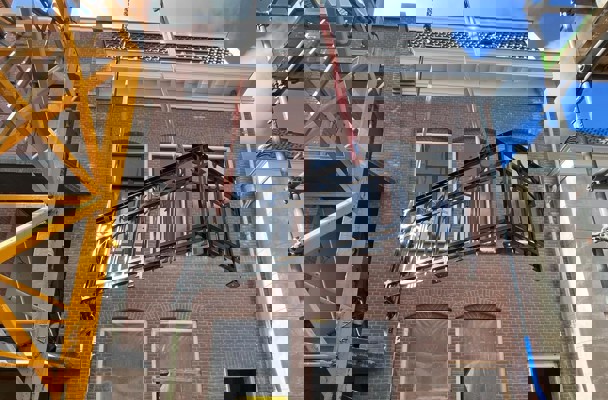 Een zwarte metalen balkonleuning wordt met een kraan omhoog getild richting een in aanbouw zijnd gebouw.