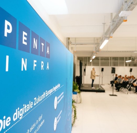 The speaker presents on a stage in front of a large audience, with a prominent blue banner of Penta Infra in the background.