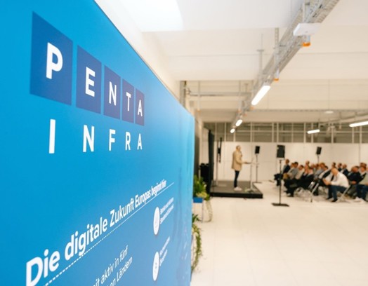 Spreker staat op een podium voor een groot publiek, met een opvallende blauwe banner van Penta Infra op de achtergrond.