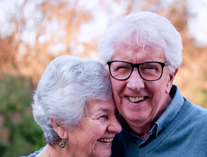 Een oudere vrouw met grijs haar en een man met grijs haar staan dicht bij elkaar en genieten van een zonnige dag in de natuur.