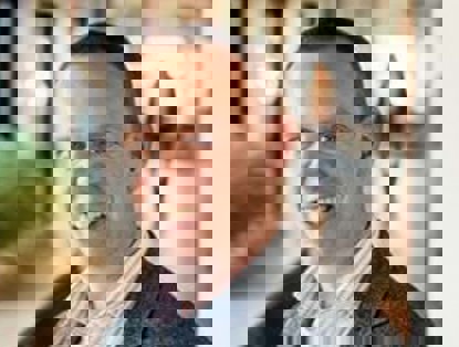 Een man in een blazer staat in een moderne, goed verlichte kantoorruimte, terwijl een vrouw op de achtergrond voorbijloopt.