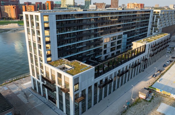 Modern square apartment complex by the water with green roofs and balconies, next to a sandy area.