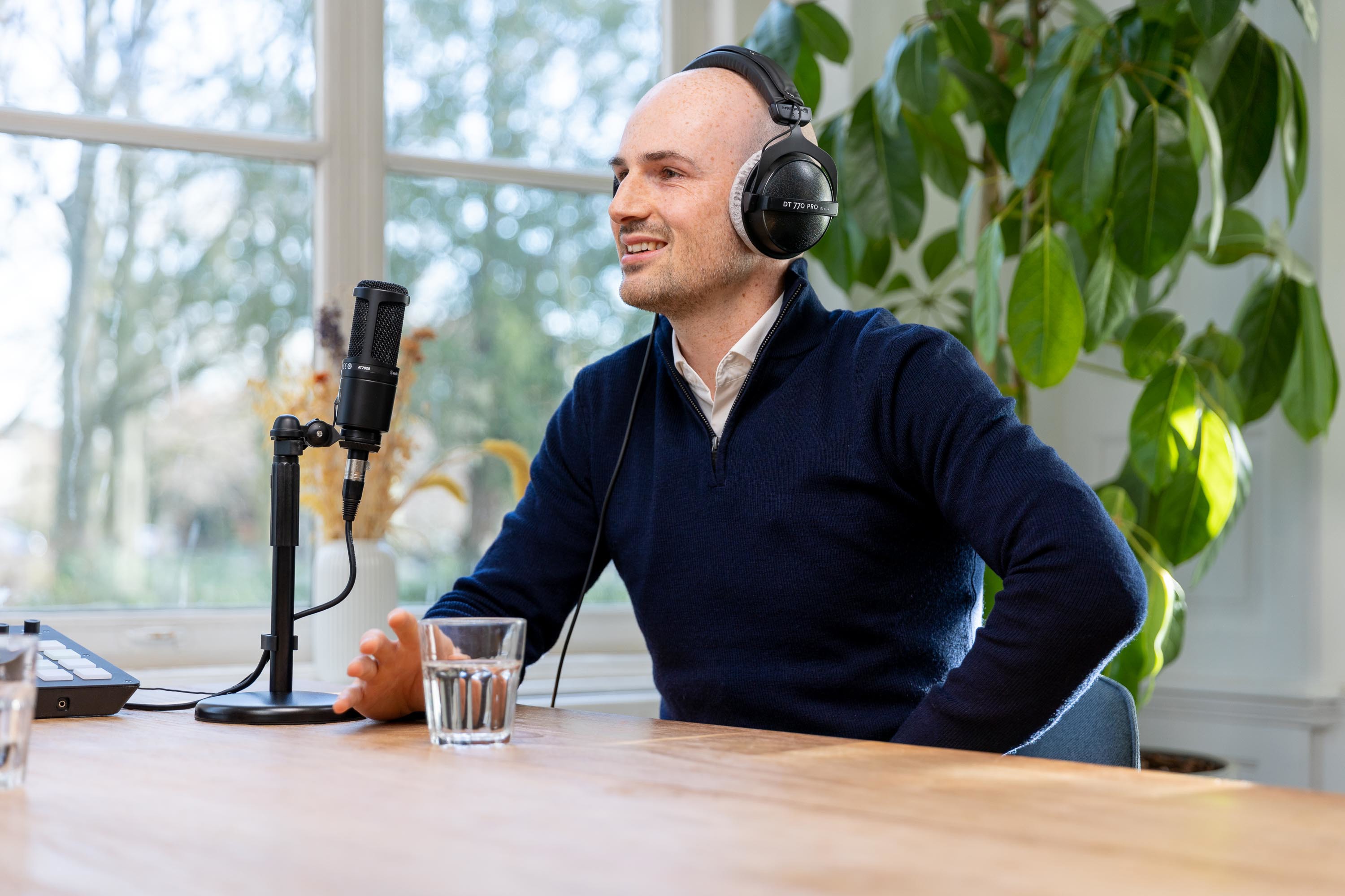 Cairan in een donkerblauwe trui, zittend aan een tafel met een microfoon en een glas water, luisterend met een headset.