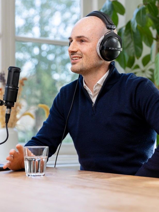 Cairan in een donkerblauwe trui, zittend aan een tafel met een microfoon en een glas water, luisterend met een headset.