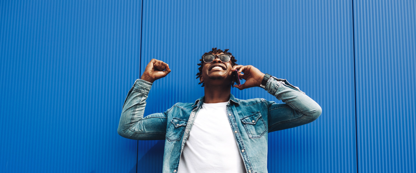 Man in een spijkerjack en wit T-shirt die met een mobiele telefoon belt, tegen een blauwe muur.