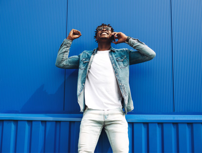 Man in een spijkerjack en wit T-shirt die met een mobiele telefoon belt, tegen een blauwe muur.