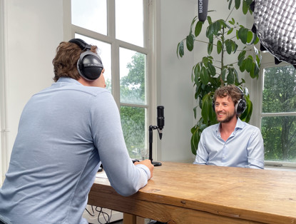 Twee personen zitten aan een houten tafel in een lichte ruimte, terwijl opnameapparatuur is opgesteld voor een gesprek.
