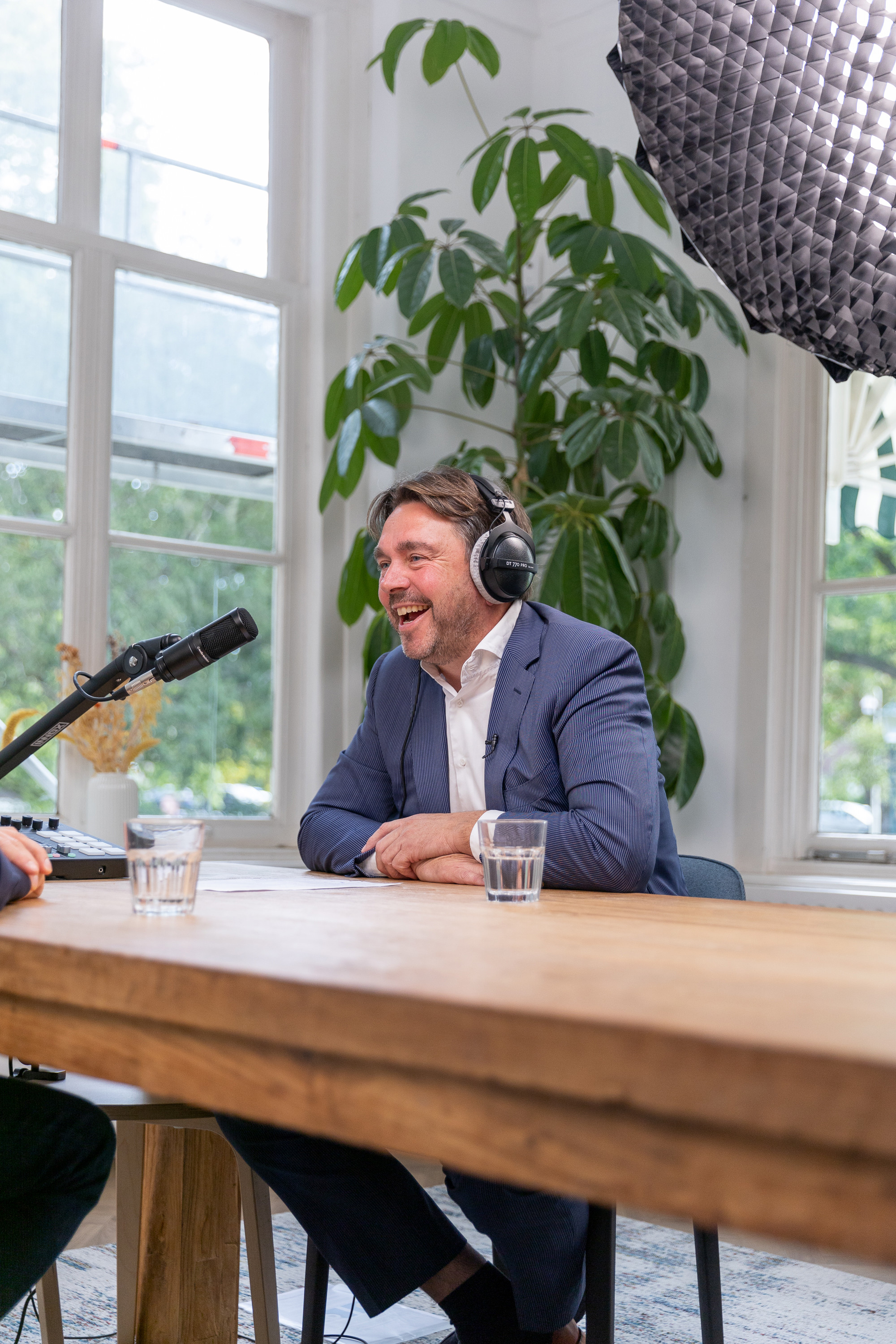 Maurits met een hoofdtelefoon zit aan een houten tafel en luistert aandachtig naar een gesprekspartner, omringd door planten en een microfoon op de achtergrond.
