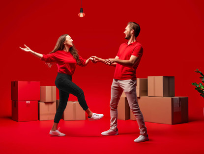 Een man en een vrouw in rode kleding dansen tegen een rode achtergrond, omringd door verhuisdozen.