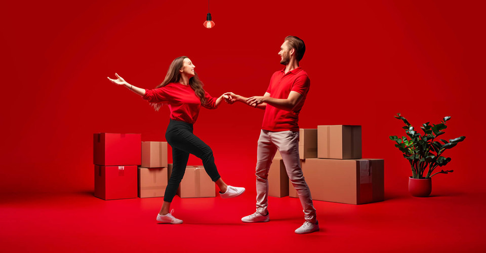 Een man en een vrouw in rode kleding dansen tegen een rode achtergrond, omringd door verhuisdozen.
