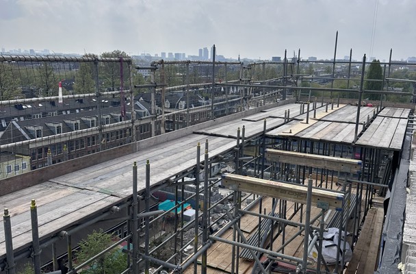 Aerial view of a construction site with steel framework and wooden planks, featuring a city skyline in the background.
