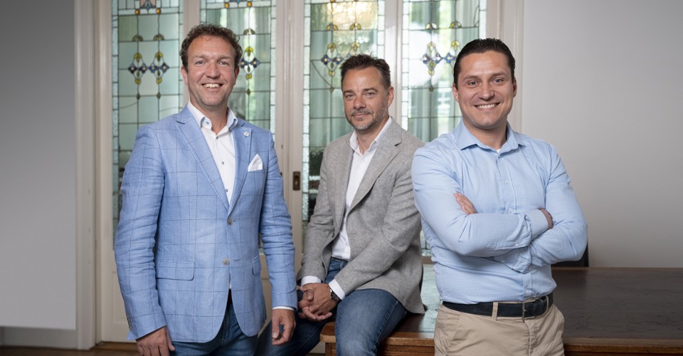 Drie mannen in zakelijke kleding staan en zitten voor een decoratieve glazen wand met loodramen.