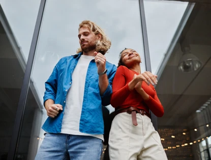 Twee mensen dansen blij voor een glazen gevel; de man draagt een blauw shirt en de vrouw een rode top.