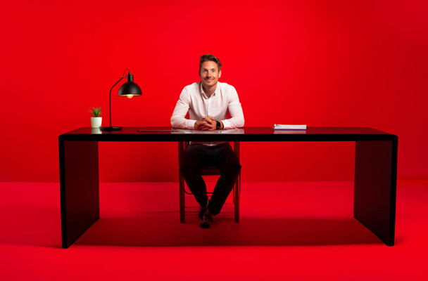 Een man in een wit shirt zit achter een zwart bureau met een tafelblad, voor een felrode achtergrond.