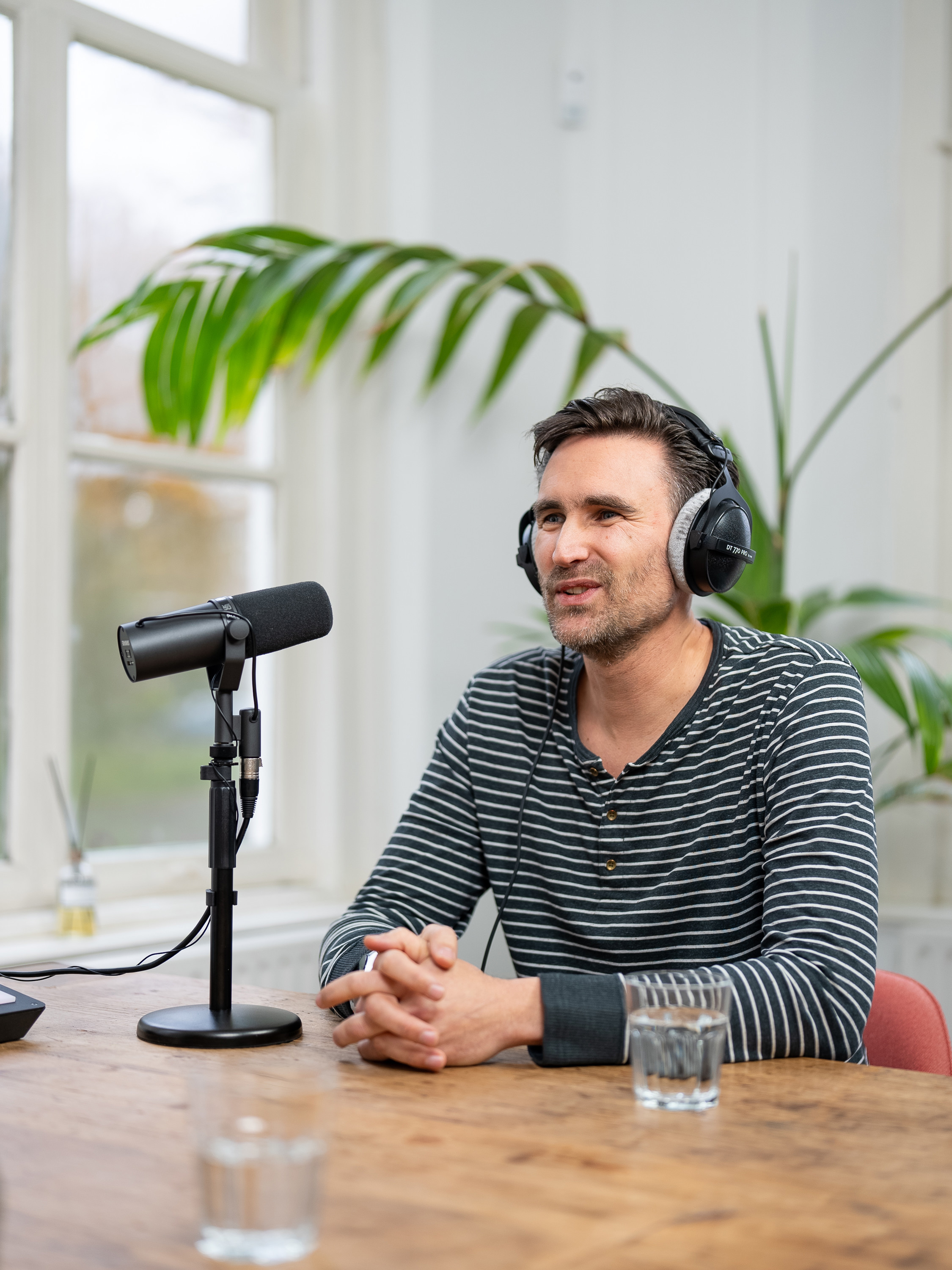 Robert met koptelefoon aan een tafel, zittend voor een microfoon in een lichte ruimte met planten op de achtergrond.