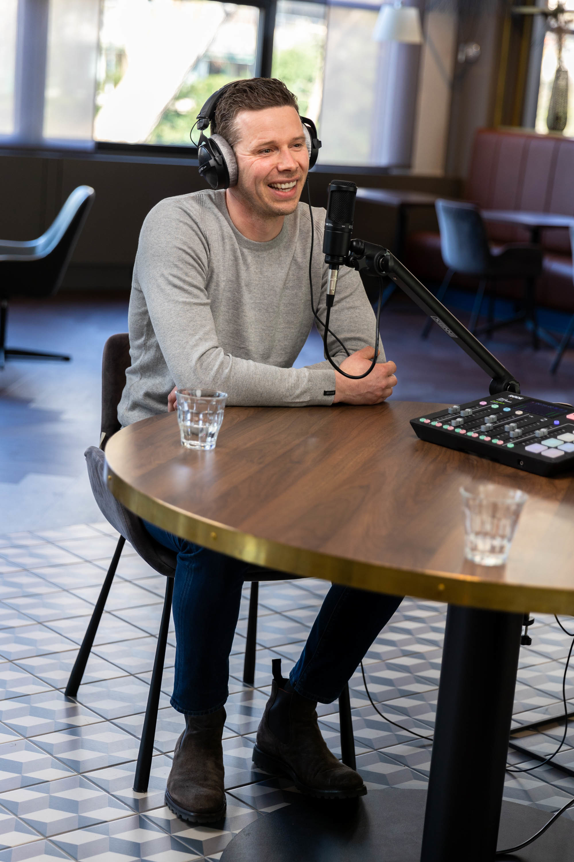 Koen in een grijs sweatshirt zit aan een ronde tafel, luisterend naar een gesprek, met een microfoon en opnameapparatuur voor zich.