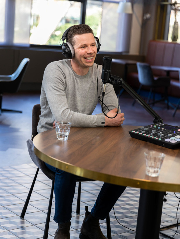 Koen in een grijs sweatshirt zit aan een ronde tafel, luisterend naar een gesprek, met een microfoon en opnameapparatuur voor zich.