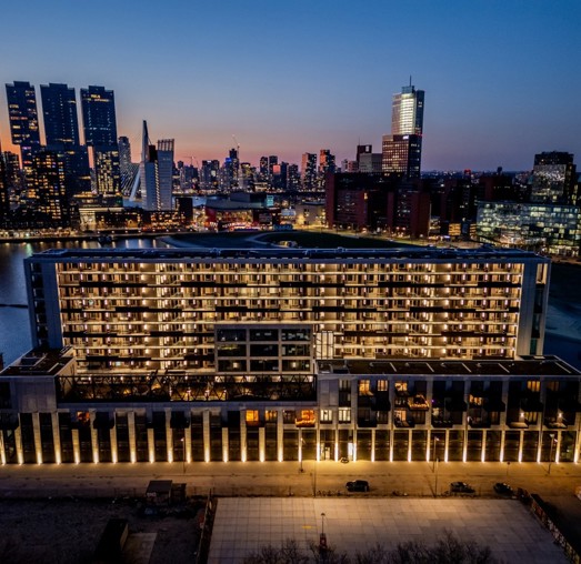 Panoramic view of a modern waterside hotel in the city, illuminated by numerous lights, with a skyline of tall buildings in the background during the evening.