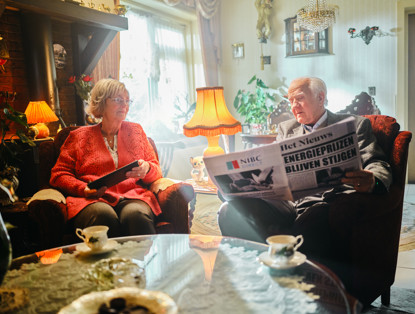 Twee ouderen zitten in een gezellige woonkamer, waarbij één van hen een krant leest met de kop 'Energietarieven blijven stijgen'.
