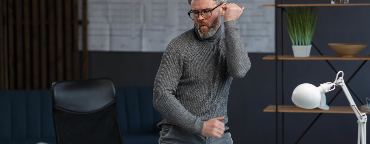 Een man in een grijze sweater danst enthousiast in een moderne kantoorruimte met een bureau en een bureaulamp.