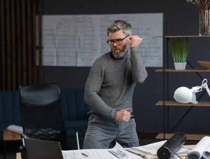 Een man in een grijze sweater danst enthousiast in een moderne kantoorruimte met een bureau en een bureaulamp.