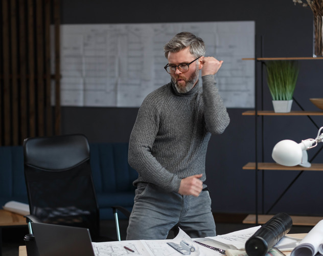 Een man in een grijze sweater danst enthousiast in een moderne kantoorruimte met een bureau en een bureaulamp.