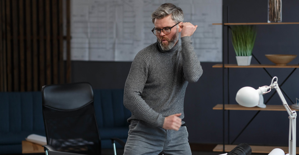 Een man in een grijze sweater danst enthousiast in een moderne kantoorruimte met een bureau en een bureaulamp.