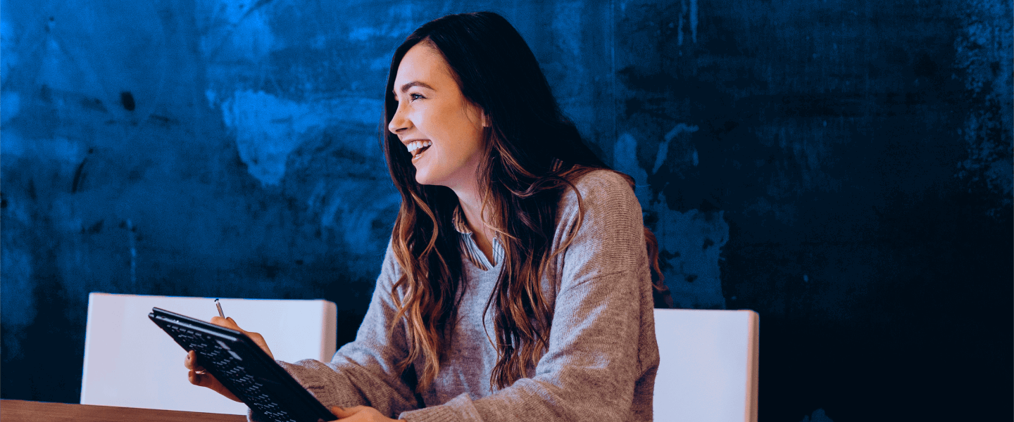 Vrouw in grijze trui zit aan een houten tafel en kijkt naar een tablet, met een blauwe muur op de achtergrond.